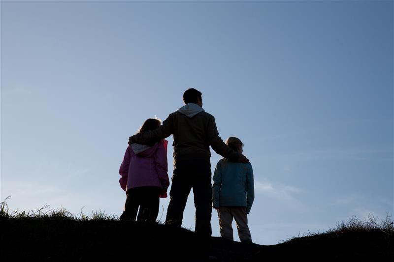 A man and two children stand with their backs to the camera, with a clear sky in the background